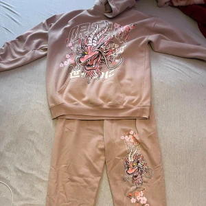 Rosa set med draktryck - Säljer ett rosa set med stort färgglatt drakmotiv och körsbärsblommor på ena benet. Både Hoodien och Byxorna är i mjukt material och har en avslappnad passform, perfekta för chill dagar. Snyggt tryck i rosa, lila och vitt som verkligen sticker ut. OBS Hoodien är i storlek S men funkar som M också, byxorna är bara S
