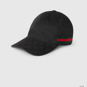 Svart keps från Gucci med logomönster - Snygg svart keps från Gucci med diskret GG-logomönster över hela ytan och klassiska gröna och röda ränder på sidan. Kepsen har böjd skärm och är tillverkad i ett slitstarkt material som ger en lyxig känsla.