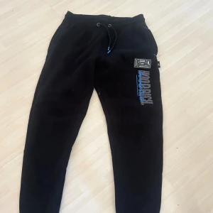 Svarta mjukisbyxor från Hoodrich - Svarta mjukisbyxor från Hoodrich med blå och vit logga på benet, snörning i midjan och muddar vid bensluten. Byxorna har tryckdetaljer både fram och bak samt en avslappnad passform. 