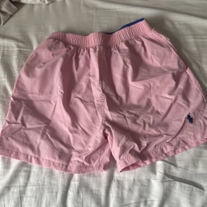 Rosa badshorts från Polo Ralph Lauren - Säljer ett par ljusrosa badshorts från Polo Ralph Lauren med elastisk midja och innerbyxa i mesh. Klassisk design med blå logga broderad på benet och bakficka med lock. Perfekta för sommardagar vid stranden eller poolen.