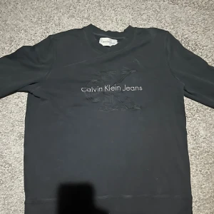 Svart sweatshirt från Calvin Klein Jeans - STÅR XXL MEN ÄR MSvart sweatshirt från Calvin Klein Jeans med broderad CK-logga och text framtill. Tröjan har rund halsringning, långa ärmar och ribbade muddar. Perfekt för dig som gillar stilrena och klassiska plagg med en modern touch.