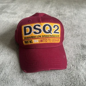 Vinröd keps från Dsquared2 - Säljer en vinröd keps från Dsquared2 med broderad patch framtill i gult, svart och vitt. Kepsen har böjd skärm och justerbar rem baktill. Tillverkad i bomull och har tydlig logga på insidan. 