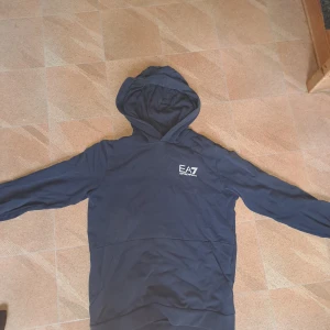 Mörkblå hoodie från EA7 Emporio Armani - Snygg mörkblå hoodie från EA7 Emporio Armani med vit logga på bröstet. Klassisk modell med huva och känguruficka framtill. Perfekt för dig som gillar stilrena och sportiga plagg. Långärmad och i mjukt material för extra komfort.