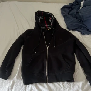 Svart Burberry hoodie med dragkedja - Svart hoodie från Burberry med klassiskt rutigt foder i huvan och insidan. Tröjan har dragkedja, snörning i huvan och broderad logga på bröstet. Mjuk och skön, perfekt för kyliga dagar. Snygg och tidlös design med två fickor framtill.