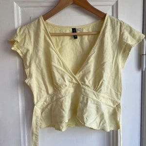 Ljusgul omlottopp från H&M Divided - Supersöt ljusgul omlottopp från H&M Divided i storlek S. Toppen har v-ringning, korta ärmar och knytband i midjan som ger en snygg passform. Tillverkad i ett lätt och luftigt material, perfekt för varma dagar.