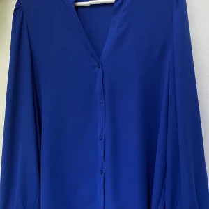 Kornblå skjorta från Vero Moda XL - Kornblå skjorta från Vero Moda i storlek XL. Skjortan har v-ringning, knappar framtill och långa ärmar med knapp vid manschetten. Tillverkad i ett lätt och mjukt polyestertyg som ger ett snyggt fall. Perfekt för dig som gillar stilrena och färgstarka plagg.