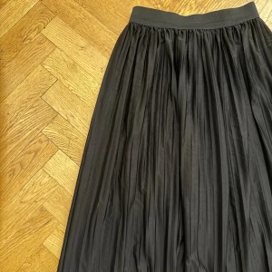 Svart plisserad kjol - Snygg svart plisserad kjol med resår i midjan. Kjolen har ett luftigt fall och slutar under knäna. Materialet känns lätt och ger kjolen ett fint flow när du rör dig. Den är i mycket fint skick!
