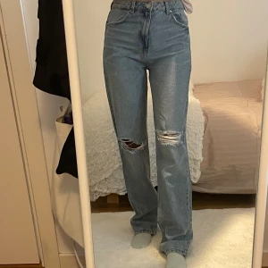 Ljusblå raka jeans med hål på knäna - Säljer ett par ljusblå jeans med raka ben och snygga slitningar över båda knäna. Jeansen har hög midja och klassisk femficksdesign. Perfekta för en avslappnad och trendig look. Materialet är klassisk denim i bomull.