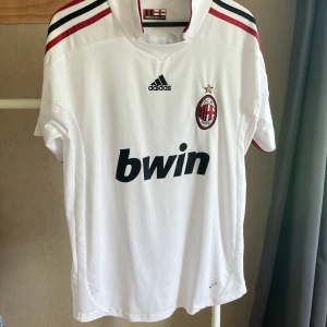 AC Milan Ronaldinho 80 Adidas tröja - Säljer en vit AC Milan fotbollströja från Adidas med Ronaldinho och nummer 80 på ryggen. Tröjan har korta ärmar, svarta och röda detaljer på axlarna, klubbmärke på bröstet och Bwin som sponsor. Materialet är lätt och andas, perfekt för match eller träning.