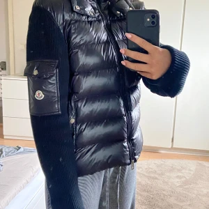 Svart Moncler Cardigan  - Säljer en svart Cardigan från Moncler med glansig dunfront och stickade ärmar. Jackan har dragkedja, hög krage, fickor med tryckknappar och Moncler-logga på ärmen. Perfekt för dig som vill ha en snygg och varm jacka.