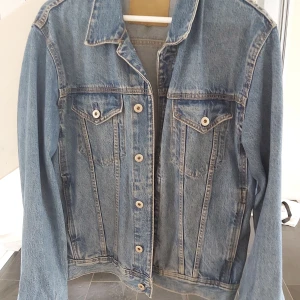Klassisk blå jeansjacka - Säljer en klassisk blå jeansjacka med knappar framtill och två bröstfickor med lock. Jackan har lång ärm och en rak passform, perfekt att slänga på sig över en hoodie eller t-shirt. Tidlös modell som funkar till det mesta.