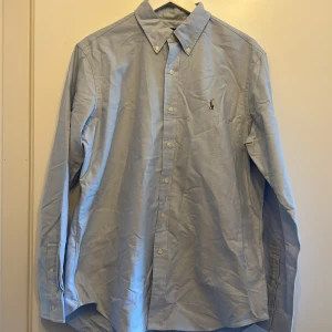 Ljusblå skjorta från Ralph Lauren - Klassisk ljusblå skjorta från Ralph Lauren med button down-krage och broderad logga på bröstet. Skjortan har långa ärmar och stängs med vita knappar framtill. Tillverkad i bomull för en skön och stilren känsla.