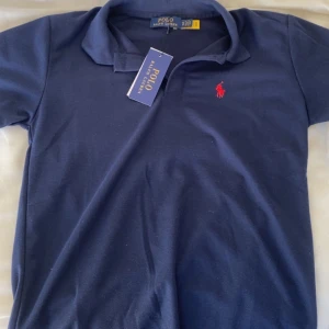 Marinblå pikétröja från Ralph Lauren - Klassisk marinblå pikétröja från Ralph Lauren med röd broderad logga på bröstet. Tröjan har krage, korta ärmar och är gjord i mjuk bomull. Perfekt för en stilren och sportig look. 