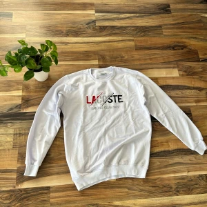 Vit sweatshirt från Lacoste M - Vit sweatshirt från Lacoste med broderad text och logga i rött, svart och grått framtill. Tröjan har rund halsringning, långa ärmar och ribbade muddar. Perfekt för en clean och sportig stil.