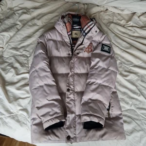Beige pufferjacka från Burberry S - MÖTS UPP I STOCKHOLM har ett litet litet hål som knappa syns, och en knapp fattas. Annars bra skick. Säljer en beige pufferjacka från Burberry med klassiskt rutigt foder och broderad logga på bröstet. Jackan har dragkedja, knappar och en patch på ärmen. Perfekt för kalla dagar och riktigt snygg med sin ikoniska design.