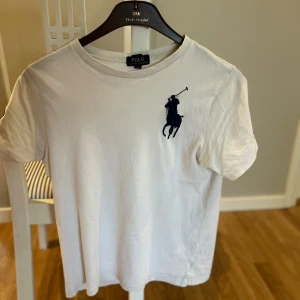 Vit t-shirt från Polo Ralph Lauren - Vit t-shirt från Polo Ralph Lauren med klassisk rund hals och stor mörkblå logga med polospelare på bröstet. T-shirten är i bomull och har en normal passform med korta ärmar. Enkel och stilren design som funkar till jeans eller shorts. Storleken är L (14-16)år han på bilden är 180cm lång. T shirten är använd 1 gång. Bara att skriva vid funderingar🙌🏼