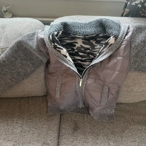 Grå pufferjacka med stickade ärmar - Snygg grå pufferjacka med stickade ärmar och ribbad krage. Jackan har dragkedja framtill, två fickor och ett mjukt foder med camouflagemönster i svart, grått och vitt. Perfekt för kyliga dagar när du vill vara både varm och stilren. Aldrig använd