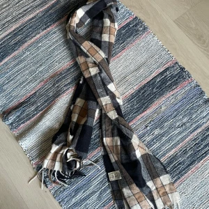 Rutig halsduk i ullmix - Snygg rutig halsduk i mjuk ullmix med fransar i ändarna. Färgkombinationen går i svart, grått, beige och vitt, vilket gör den lätt att matcha till olika outfits. Perfekt accessoar för kyliga dagar och ger en stilren touch till din look. Har glömt bort vilket märke, men köptes på Åhléns.
