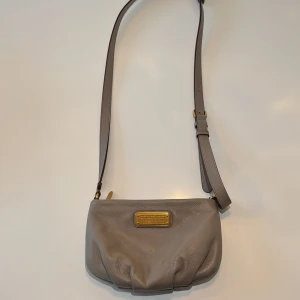 Grå axelväska Marc by Marc Jacobs - Snygg trendig grå axelväska från Marc by Marc Jacobs i mjukt skinn. Väskan har justerbar rem, guldfärgad metallplatta framtill och dragkedja upptill. Perfekt storlek för det viktigaste och passar till många olika outfits. Väska som är i nyskick!