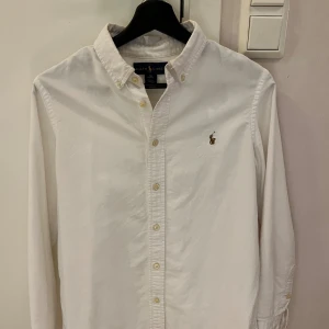 Vit skjorta från Ralph Lauren - Klassisk vit skjorta från Ralph Lauren med broderad logga på bröstet. Skjortan har lång ärm, button down-krage och knäppning framtill. Snygg custom fit-modell i bomull som passar perfekt till jeans eller chinos. Storleken är 16(år) samma som storlek (S) herr