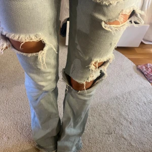 Ljusblå slitna bootcut jeans - Säljer ett par ljusblå jeans med flera stora slitningar och hål över knäna och låren. Jeansen har bootcut-modell med lätt utsvängda ben och klassisk femficksdesign. Perfekta för en avslappnad och trendig look.