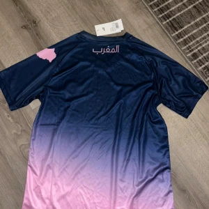 Marocko fotbollströja Puma blå/rosa - Snygg Marocko fotbollströja från Puma i ett glansigt material. Tröjan har en cool blå till rosa färgövergång, korta ärmar och tryck i rosa med arabisk text och karta. Perfekt för dig som vill sticka ut på match eller träning.