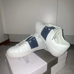 Valentino Garavani vita sneakers - Snygga vita sneakers från Valentino Garavani med bred marinblå detalj över sidan och klassiska nitar på hälen. Skorna har snörning och är tillverkade i skinn för en lyxig känsla. Perfekta för dig som vill ha en stilren men ändå unik look.