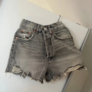 Gråa jeansshorts med fransad kant från Zara - Snygga grå jeansshorts från Zara med hög midja och fransad, sliten kant. Shortsen har klassiska fem fickor, knapp och dragkedja framtill samt coola slitningar både fram och bak. Perfekta för dig som gillar en avslappnad och trendig stil.