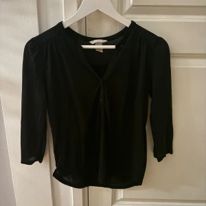 Svart blus från H&M med knappar XS - Svart blus från H&M i storlek XS. Modellen har trekvartsärm, v-ringning och knappar framtill. Tunt och lätt material som ger en avslappnad känsla. Perfekt att styla med jeans eller kjol för en enkel men snygg look.