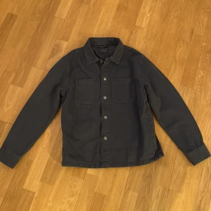 Svart overshirt från No Boundaries M - Säljer en svart overshirt från No Boundaries i bomull. Jackan har två stora bröstfickor, knappar framtill och klassisk krage. Perfekt att slänga på sig över en hoodie eller t-shirt. Enkel och stilren design med lång ärm och snygga detaljer vid ärmslut. Storlek M, säljer för att den inte passar längre. Bara använt ett par gånger.
