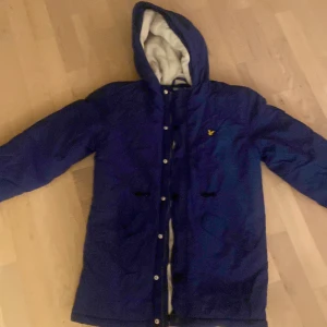 Blå parkajacka från Lyle & Scott ( kan visa mer bilder) - Mörkblå parkajacka från Lyle & Scott med huva och mysigt vitt fleecefoder. Jackan har knappar framtill, klassisk gul logga på bröstet och är perfekt för kyliga dagar. Snygg och funktionell design som håller dig varm hela vintern.