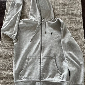 Grå hoodie från Polo Ralph Lauren - Snygg ljusgrå hoodie från Polo Ralph Lauren med dragkedja framtill och klassisk huva med snörning. Tröjan är sparsamt använd, som ny.