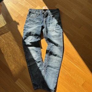 SMOG blå jeans med slitningar - Klassiska SMOG jeans i blå tvätt med snygga slitningar och kontrastsömmar. Rak passform, fem fickor och knappgylf. Jeansen har en autentisk vintagekänsla och är tillverkade i robust denim. Perfekta för dig som gillar en avslappnad och tidlös stil.
