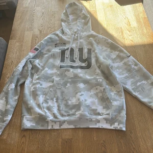 NY Giants camo hoodie NFL - Säljer en grå och vit camo-mönstrad hoodie från NFL med stor NY Giants-logga på bröstet. Tröjan har huva med snörning, känguruficka och amerikansk flagga på ärmen. Perfekt för dig som gillar sportig streetstyle och vill sticka ut.
