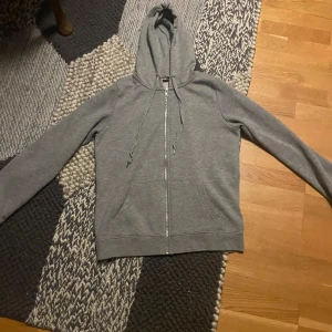 Grå hoodie från FB Sister med dragkedja - En stilren grå hoodie från FB Sister i storlek S. Tröjan har huva med snörning, hel dragkedja framtill och två fickor. Tillverkad i mjukt material, perfekt för chill dagar eller när du vill ha en enkel och clean look.