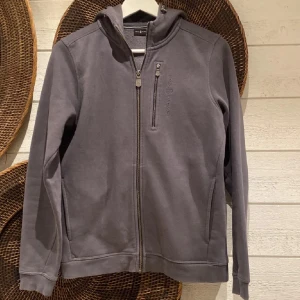 Grå hoodie från Sail Racing - Säljer en grå hoodie från Sail Racing med dragkedja framtill och bröstficka med dragkedja. Tröjan har huva med snörning och broderad logga på bröstet. Mjuk insida och ribbade muddar vid ärmslut och nederkant. Perfekt för kyliga dagar.