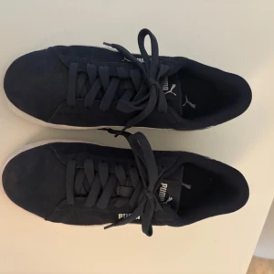 Mörkblå sneakers från Puma - Snygga mörkblå sneakers från Puma med klassisk vit sula och vit sidodetalj. Skorna har rund tå, platt sula och snörning. Ovandelen är i mocka för en stilren och sportig look. Perfekta för dig som gillar streetstyle och enkel design.