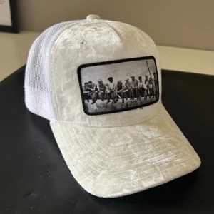 Vit sammetstruckerkeps med fotoapplikation - Snygg vit truckerkeps i glansig sammet med nät baktill. Framsidan har en stor svartvit applikation med det ikoniska fotot 'Lunch atop a Skyscraper'. Justerbar baktill för perfekt passform.
