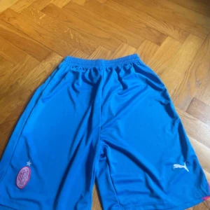 Blå AC Milan Puma shorts - Säljer ett par blå sportiga shorts från Puma med AC Milan-logga på ena benet. De har elastisk midja och är tillverkade i ett lätt och svalt material som passar perfekt för fotboll eller träning.