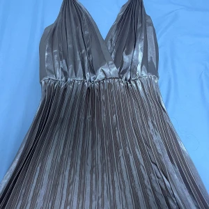 Champagnefärgad plisserad långklänning - Säljer en elegant långklänning i champagnefärgat, glansigt tyg med omlott-v-ringning och breda axelband. Klänningen har plisserad kjol och smockad rygg för extra komfort. Perfekt för dig som vill sticka ut med en lyxig och stilren look.