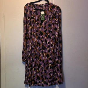Lila mönstrad klänning från Indiska - Säljer en snygg kortklänning från Indiska med abstrakt mönster i lila, svart, brun och beige. Klänningen har lång ärm och rund halsringning. Tillverkad i mjuk polyester som faller fint. Perfekt för dig som gillar färg och mönster!