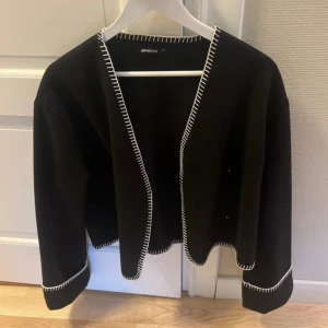 Svart kofta/jacka med vita detaljer - Svart kofta/jacka från Gina Tricot med dekorativa vita sömmar längs kanterna och ärmsluten. 