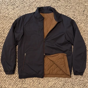 Loro Piana jacka  - Snygg vändbar jacka med ena sidan i mörkblått och andra i brun, båda med quiltad design. Jackan har dragkedja, två fickor med dragkedja och elastiska muddar. Perfekt för dig som vill variera din stil mellan klassiskt marinblått och trendigt brunt.