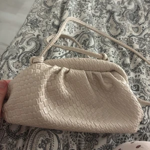 Beige axelväska i croco-imitation - Snygg beige clutch med croco-imitation och mjuk, rundad form. Väskan har ett smalt axelband och är tillverkad i syntetmaterial med struktur som påminner om krokodilskinn. Perfekt för dig som vill ha en trendig och stilren accessoar.