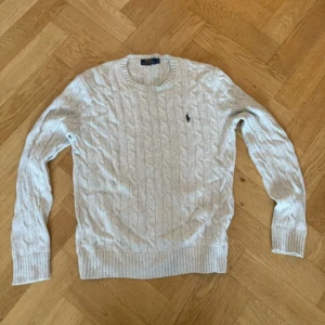 Ljusgrå kabelstickad tröja Polo Ralph Lauren - Kabelstickad ljusgrå tröja från Polo Ralph Lauren i 100% bomull. Klassisk rund halsringning, ribbade muddar och ikonisk broderad logga på bröstet. Perfekt för lager-på-lager och snygg till jeans