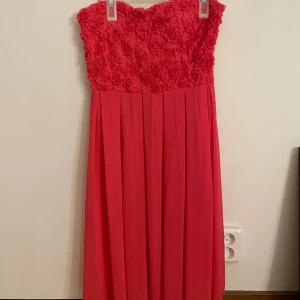 Röd axelbandslös långklänning med rosor - Snygg röd axelbandslös långklänning med 3D-blommor på överdelen och plisserad kjol. Klänningen har en romantisk vibe med rosdetaljer och en flowy känsla. Perfekt för dig som vill sticka ut med en färgstark och elegant look.