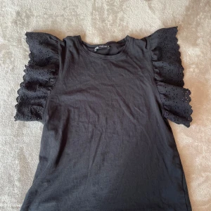 Svart blus med volangärmar från Zara - Supersnygg svart blus från Zara med breda volangärmar i broderad bomull. Ärmarna har ett blommigt hålmönster och ger en cool statement-look. Rund halsringning och rak passform. Perfekt att styla med jeans eller kjol för en trendig vibe.
