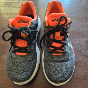 Asics sneakers svart/orange - Säljer ett par Asics sneakers i svart och orange med silvriga detaljer. Skorna har snörning, ventilerande mesh-ovandel. Snygg sportig look och bekväm passform, perfekta för träning eller vardagsstil. Är använda en del men har fortfarande mycket att ge.