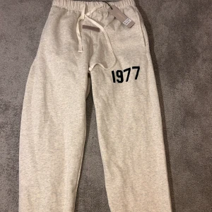 Essentials Fear of God mjukisbyxor grå M - Grå mjukisbyxor från Essentials Fear of God med trycket '1977' på benet. Byxorna har elastisk midja med vit snörning och mjukt material i bomullsmix. Perfekt för chill dagar och streetwear-stil.
