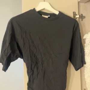 Svart t-shirt med knyt från Nelly - Svart t-shirt från Nelly i storlek XXS. Modellen har korta ärmar, rund halsringning och en snygg knyt-detalj i ryggen som ger en cool twist. Tillverkad i mjuk bomull för en skön känsla. Perfekt för dig som gillar stilrena basplagg med det lilla extra.